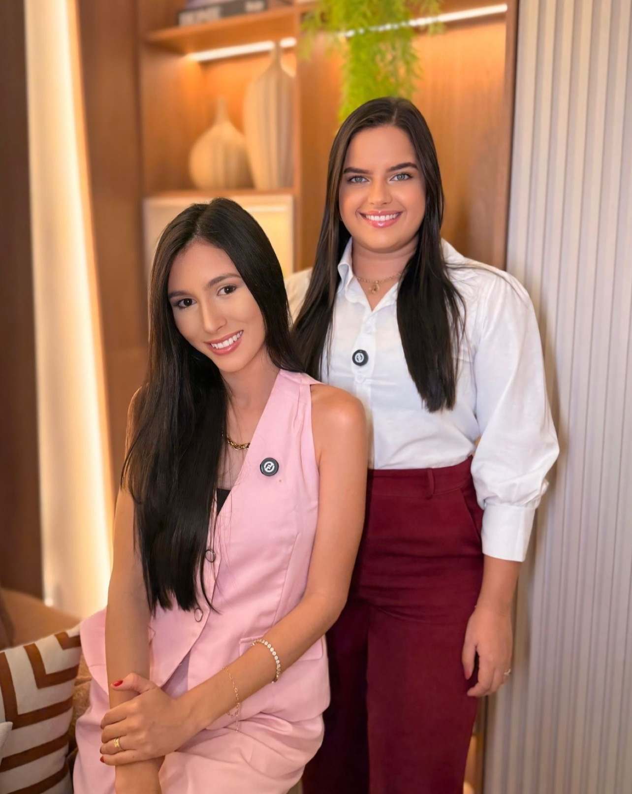 Mariana e Jéssica, sócias da Marje Arquitetura e Interiores, lado a lado diante de um nicho de madeira iluminado.